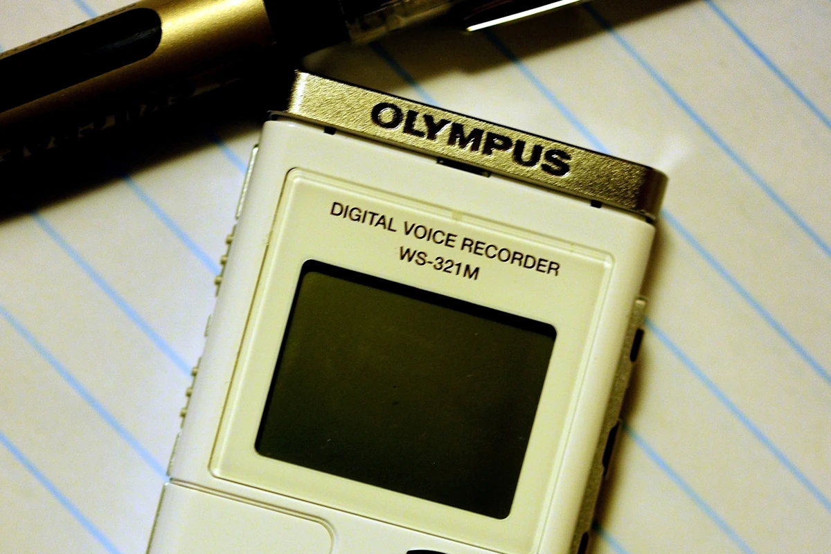 A small white Olympus digital voice recorder lying on lined note paper beside a pen